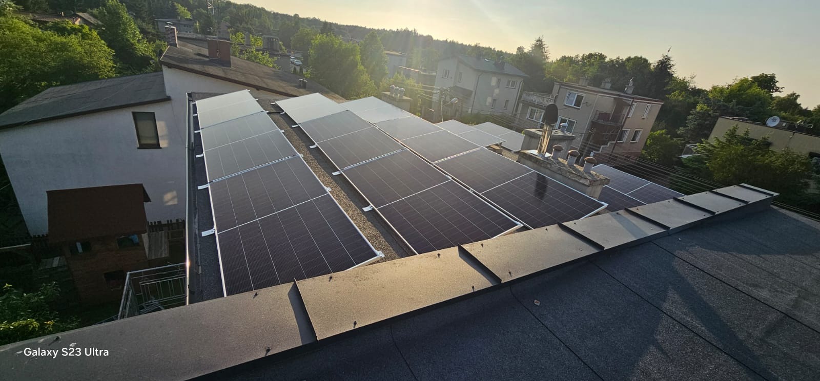 Instalacja fotowoltaiczna na dachu budynku mieszkalnego, widok z góry, panele w układzie rzędowym, w tle zieleń drzew i zabudowa.