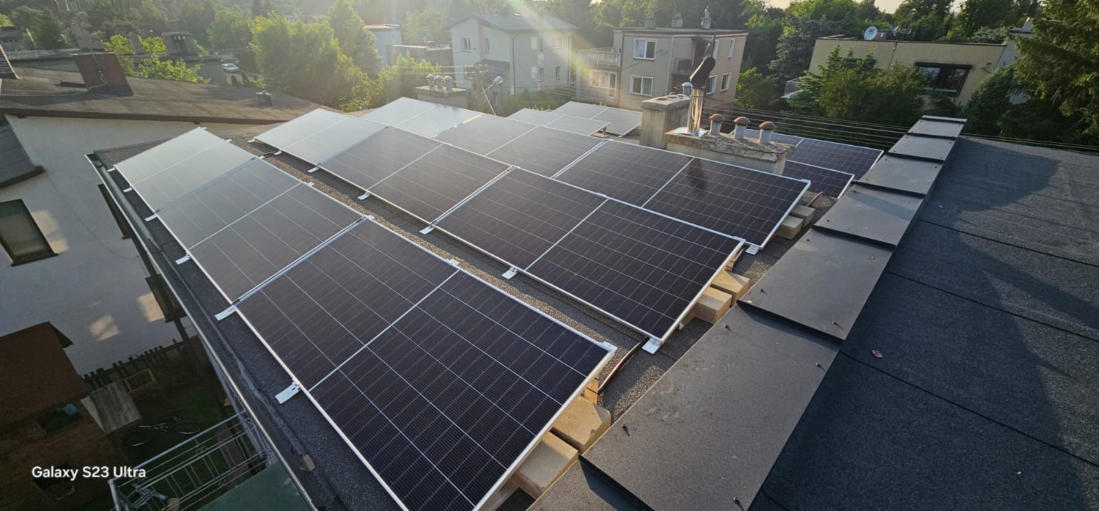 Panele fotowoltaiczne na dachu budynku mieszkalnego, zamontowane na specjalnych podkładkach, widok z góry, w tle zabudowa miejska i zieleń.
