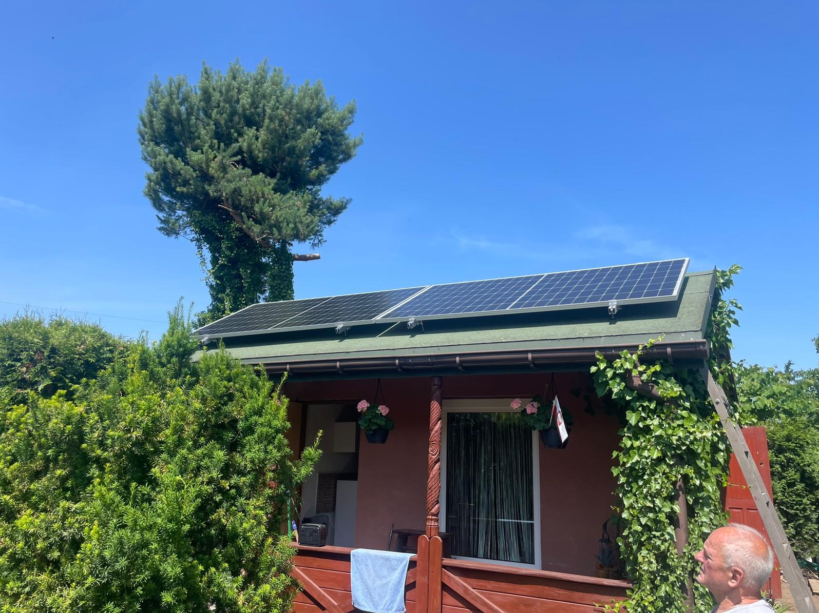 Panele fotowoltaiczne na dachu domku letniskowego z zielonym dachem, na tle błękitnego nieba i drzew. Instalacja fotowoltaiczna na małej architekturze.