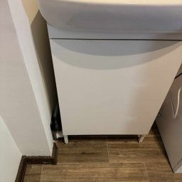 Oleg Siedykh - Biała umywalka z szafką w łazience, widok z dołu, z drewnianą podłogą i listwą przypodłogową. Ujęcie na zainstalowaną umywalkę z bliska.