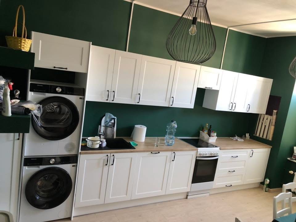 Biała zabudowa kuchenna z pralką i suszarką w kolumnie. Nowoczesne wnętrze z zielonymi ścianami i lampą wiszącą. Widok na blat z akcesoriami kuchennymi.