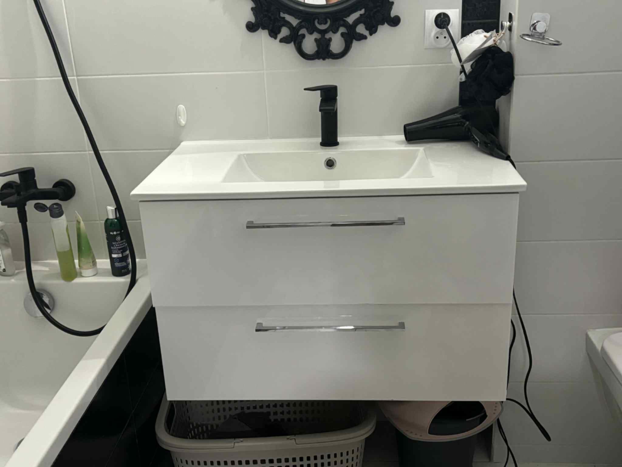 Biała szafka pod umywalkę z czarną baterią w nowoczesnej łazience z czarno-białymi płytkami. Suszarka do włosów leży na blacie, a pod szafką kosz na pranie.