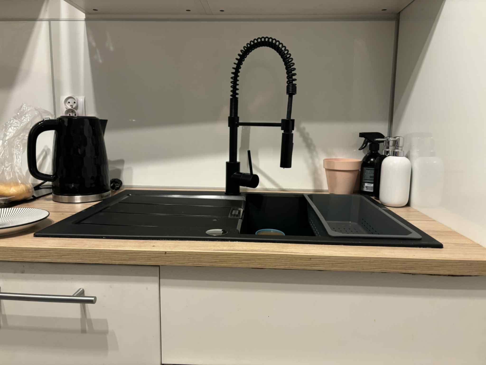 Czarny, nowoczesny kran z elastyczną wylewką nad czarnym zlewozmywakiem w jasnej kuchni. Widoczne akcesoria kuchenne i gniazdko elektryczne.