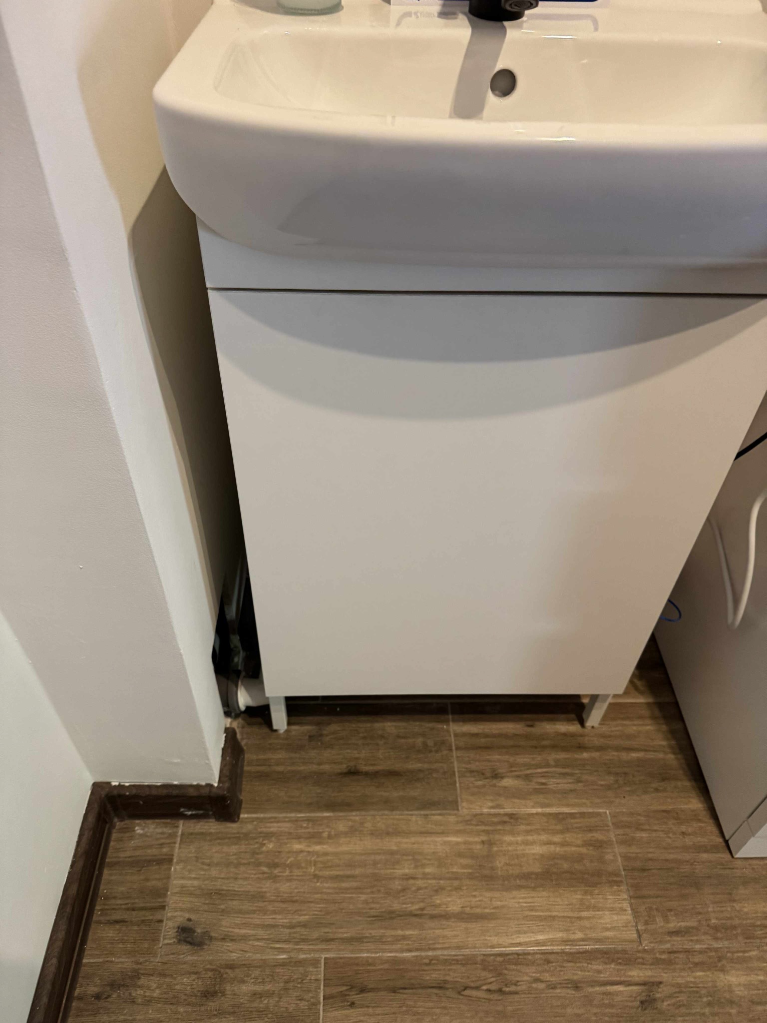 Biała umywalka z szafką w łazience, widok z dołu, z drewnianą podłogą i listwą przypodłogową. Ujęcie na zainstalowaną umywalkę z bliska.