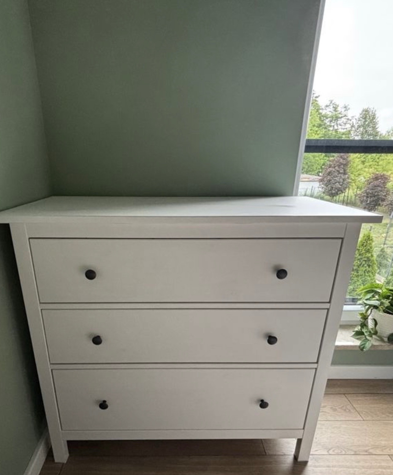 Biała komoda z trzema szufladami, umieszczona w rogu pokoju z oknem, przez które widać zieleń. Minimalistyczny design, czarne okrągłe uchwyty. Jasna podłoga.