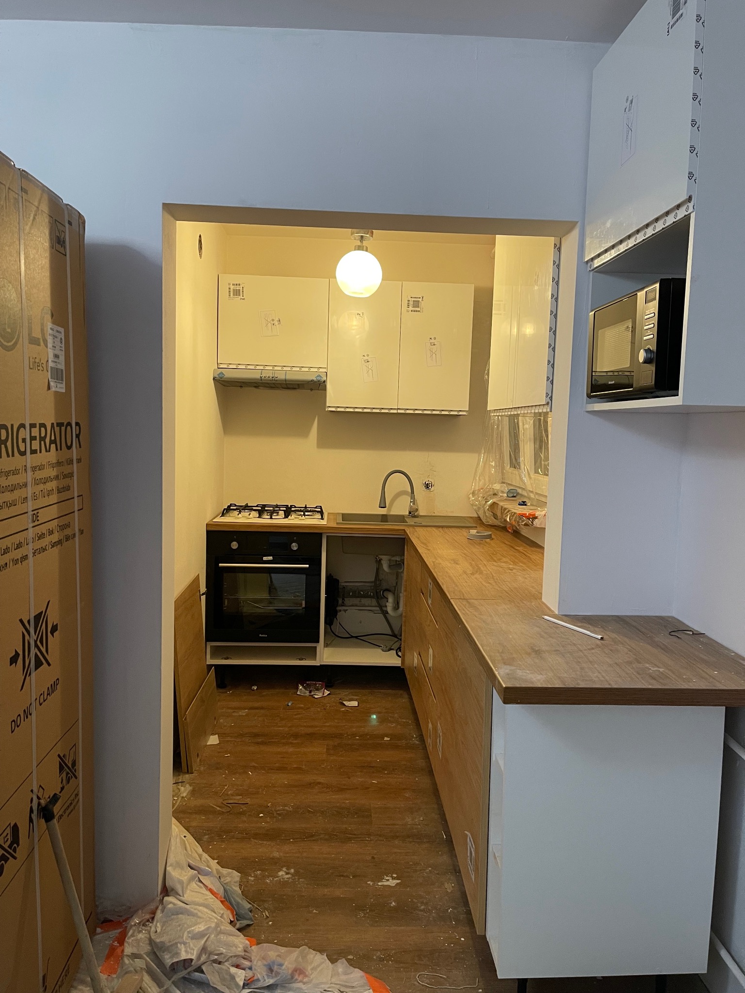 Wnętrze kuchni w trakcie montażu: widoczne szafki wiszące, kuchenka, piekarnik, drewniany blat i kartonowe pudło lodówki. Częściowy montaż zabudowy kuchennej.