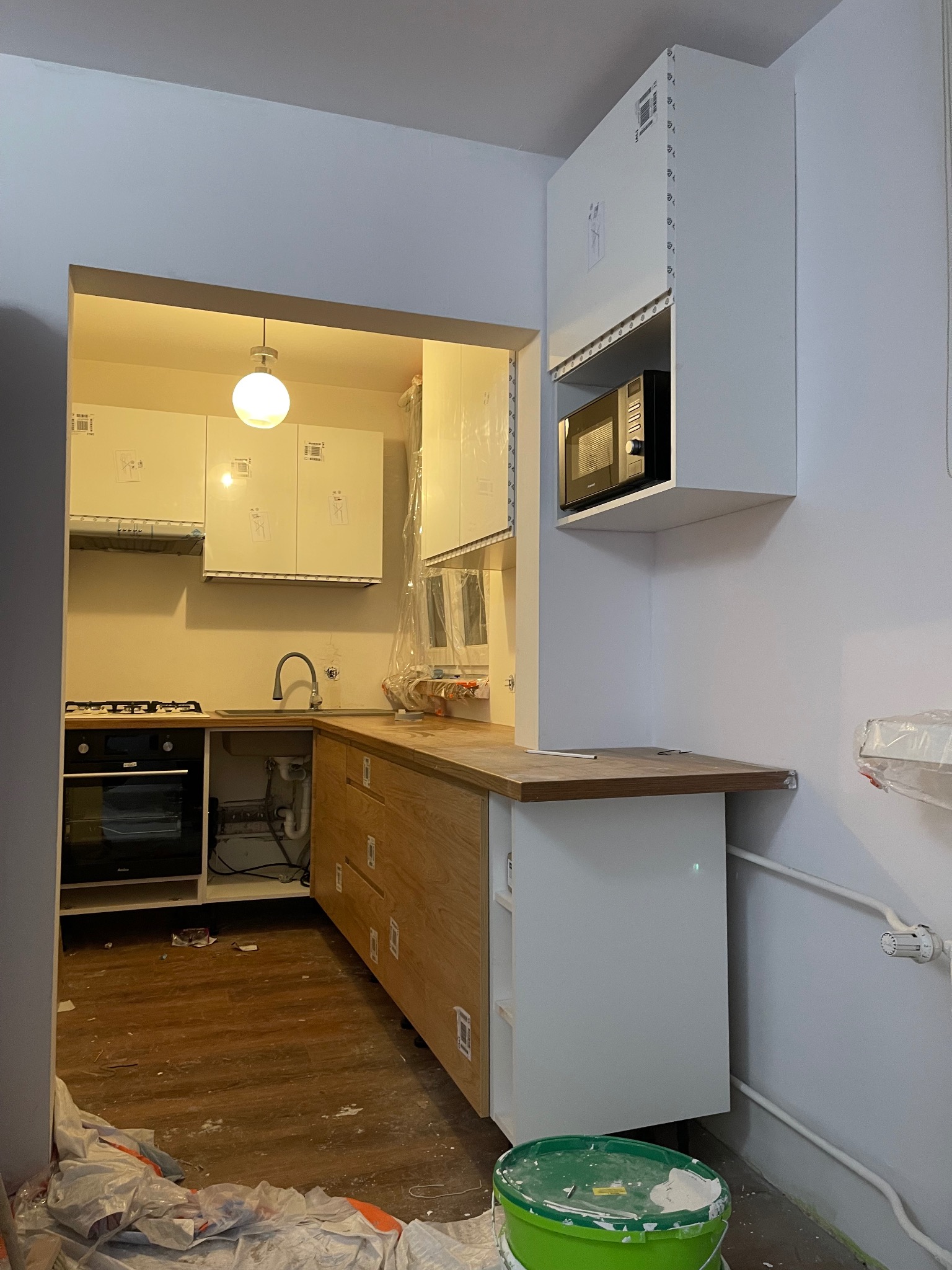 Wnętrze kuchni w trakcie montażu: białe szafki górne i dolne z drewnianymi frontami szuflad, kuchenka, zlew. Na podłodze folia malarska i wiadro z farbą.