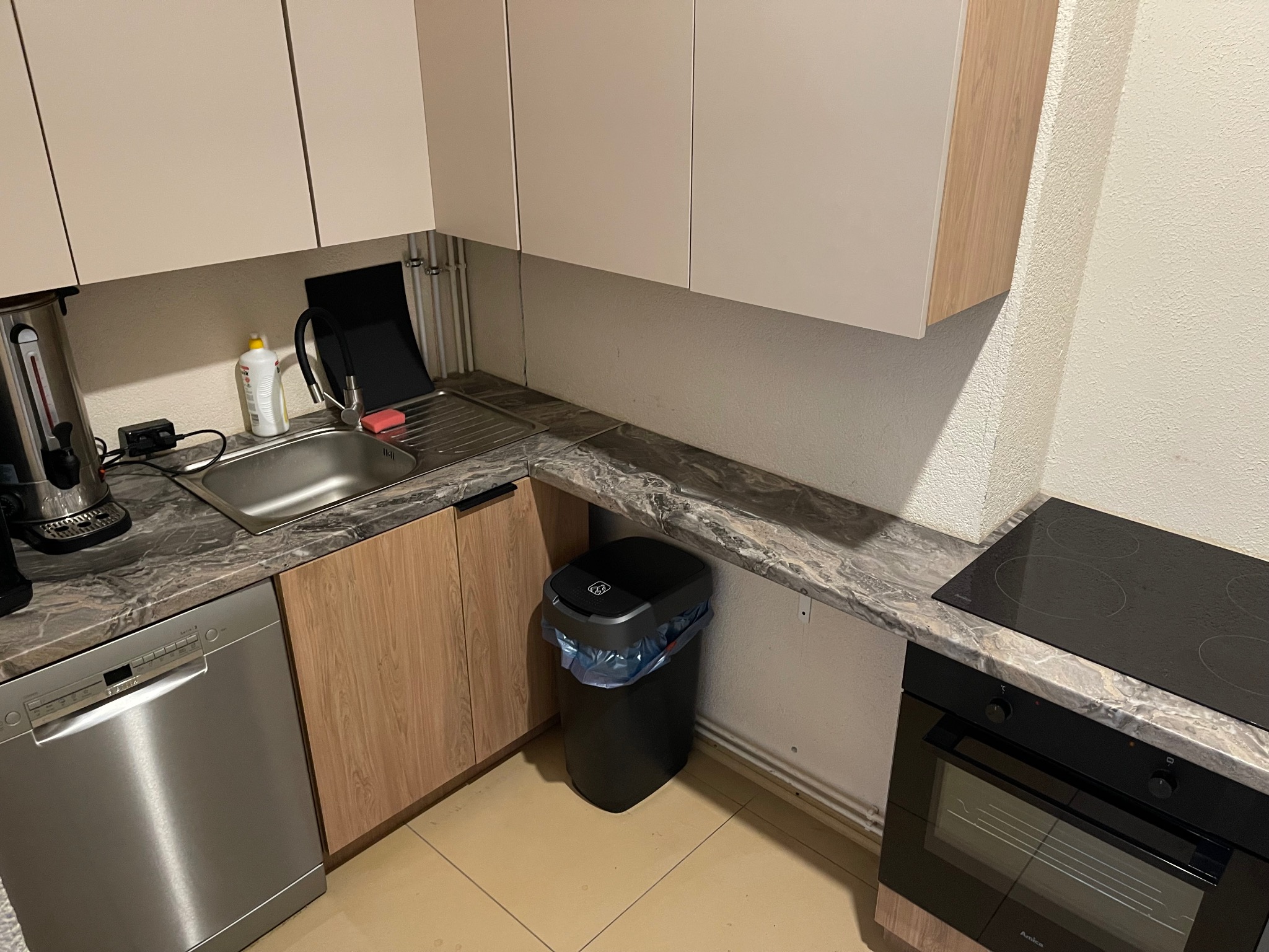 Kuchnia z zabudową, szary blat imitujący marmur, srebrna zmywarka, czarny kosz na śmieci i piekarnik z płytą indukcyjną. Jasne płytki na podłodze.