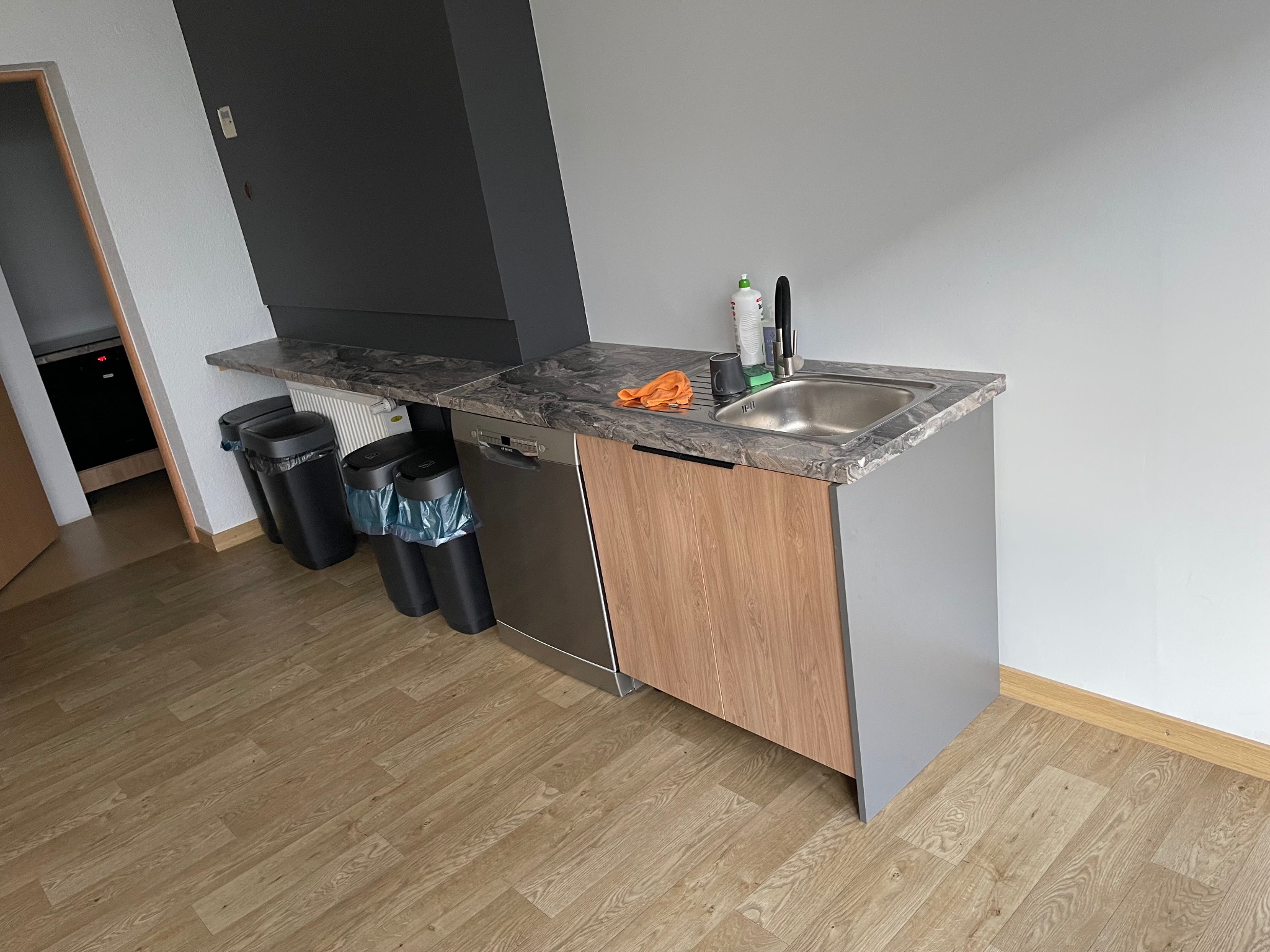 Kompaktowa kuchnia z szarą zabudową i marmurowym blatem, zlewem, baterią i pomarańczową ścierką. Obok zmywarka i kosze na śmieci. Ujęcie z niskiego kąta.