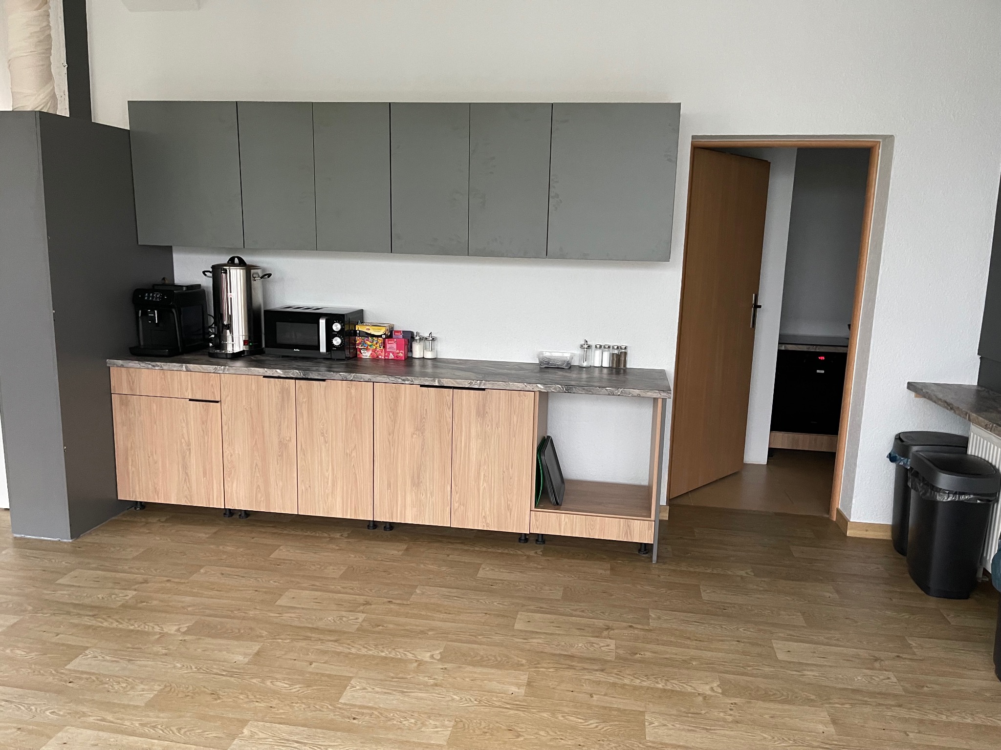 Jasna kuchnia biurowa z drewnianymi szafkami i szarymi szafkami wiszącymi. Na blacie ekspres do kawy, czajnik i mikrofalówka. Na podłodze panele imitujące drewno.
