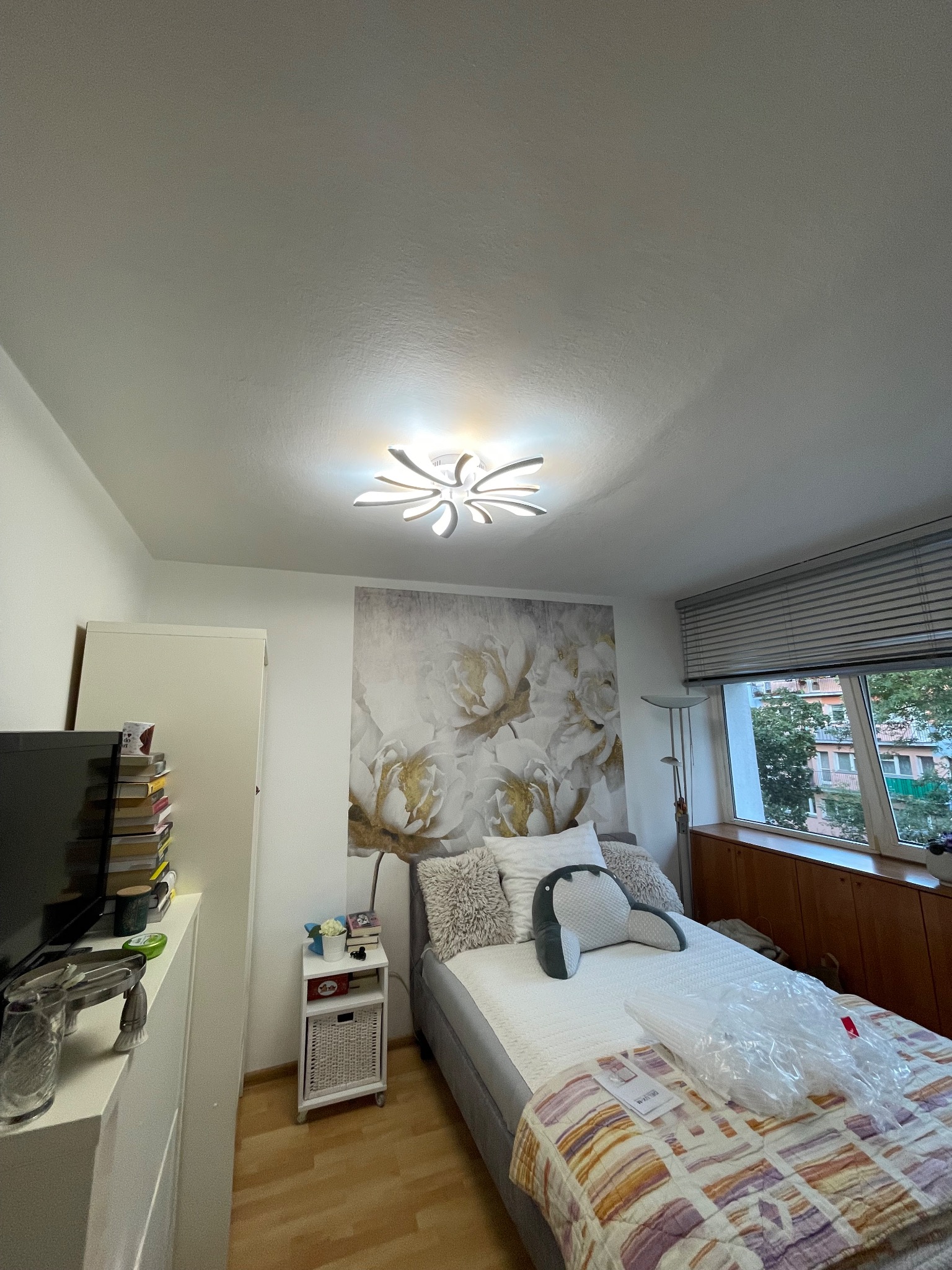 Nowoczesna lampa sufitowa w sypialni z łóżkiem i kwiatową tapetą. Lampa oświetla pomieszczenie, tworząc przytulną atmosferę. Widok na okno z żaluzjami.