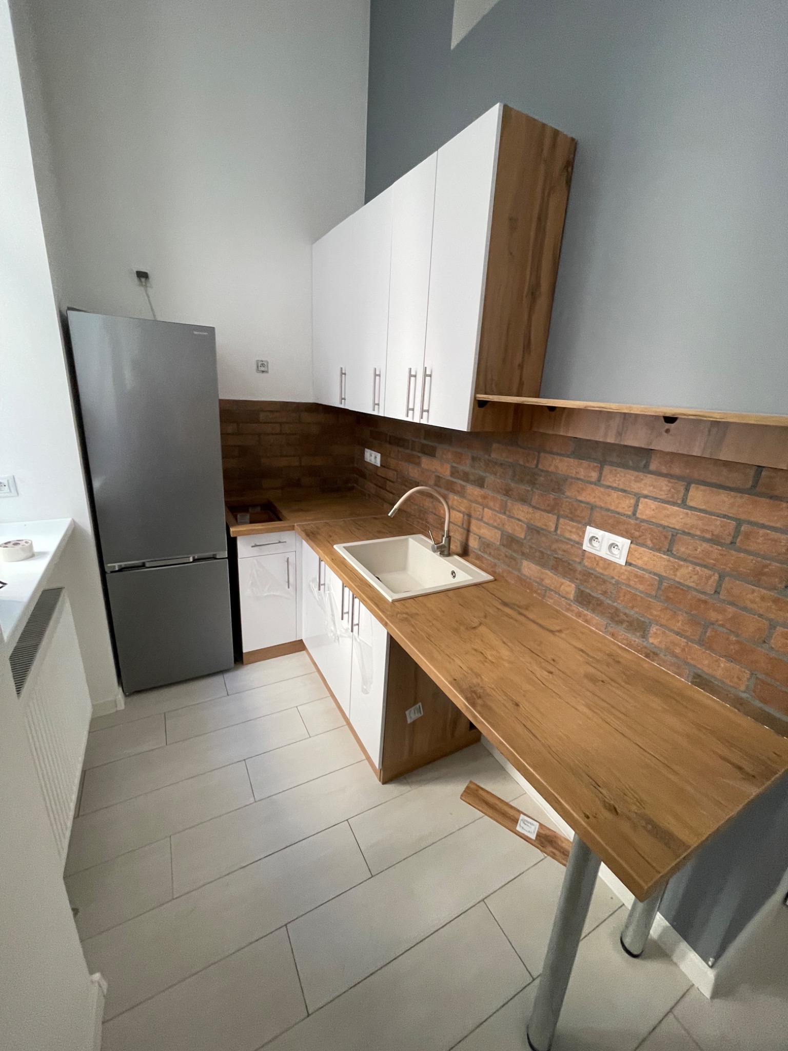 Nowoczesna kuchnia z białymi szafkami, drewnianym blatem i ceglaną ścianą. Uwagę przykuwa wysuwany blat stołu wsparty na metalowej nodze.