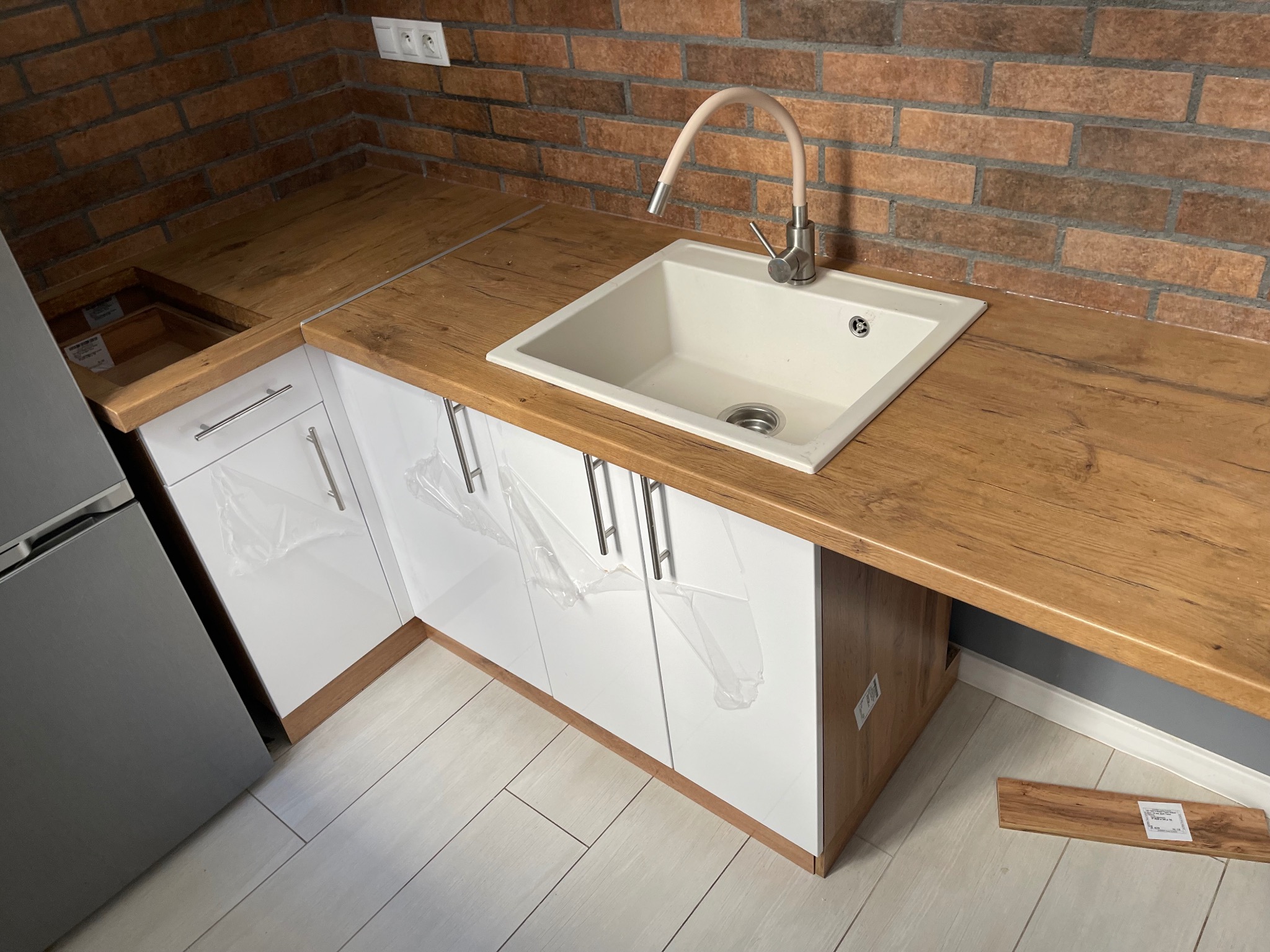 Nowoczesna kuchnia z białymi szafkami, drewnianym blatem i ceglaną ścianą. Umywalka z baterią. Częściowo zdjęta folia ochronna z szafek. Widoczny fragment lodówki.