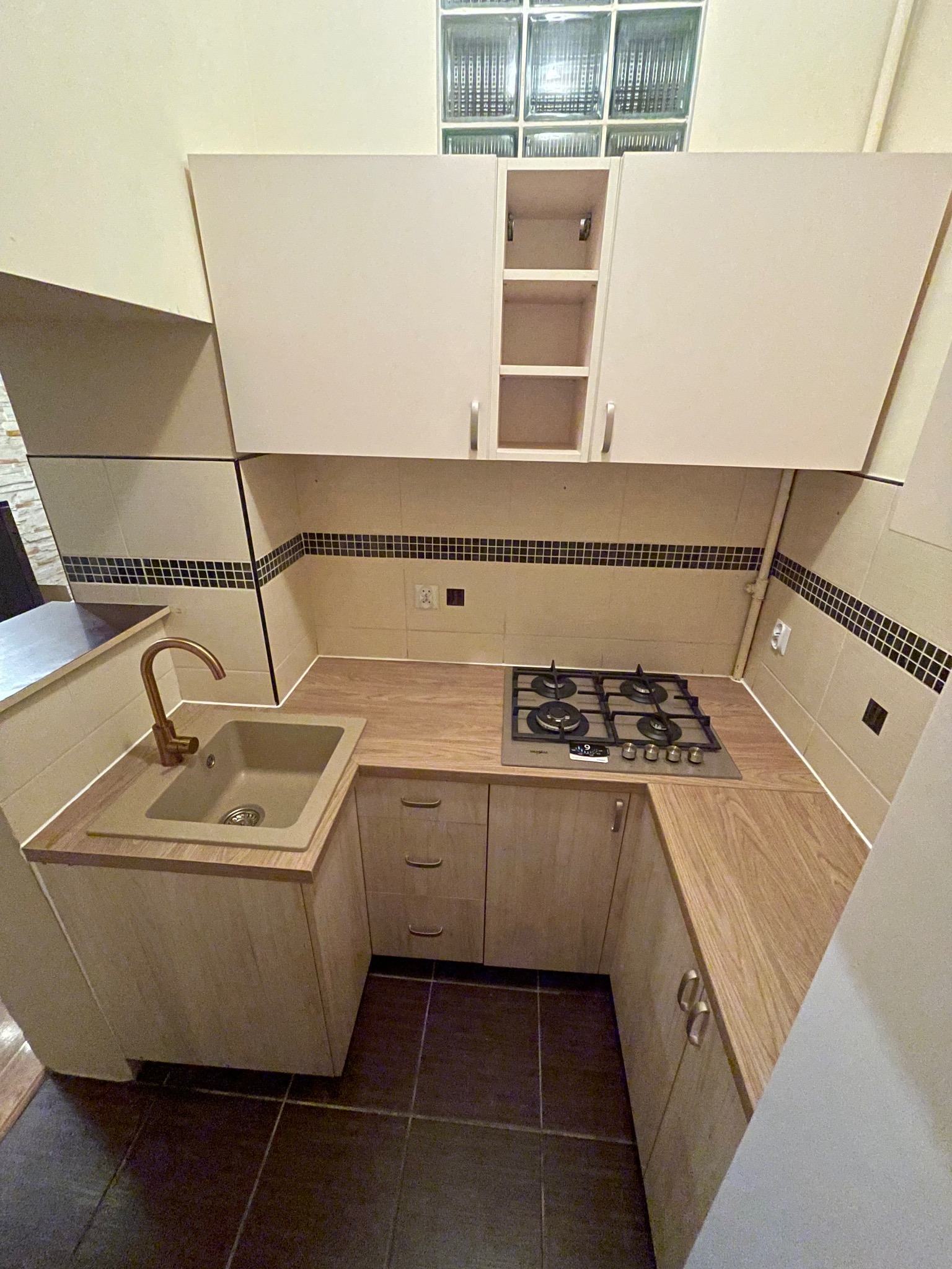 Jasna kuchnia w bloku po remoncie: beżowe szafki z drewnianym blatem, złota bateria przy zlewie i czarna płyta gazowa. U góry luksfery.