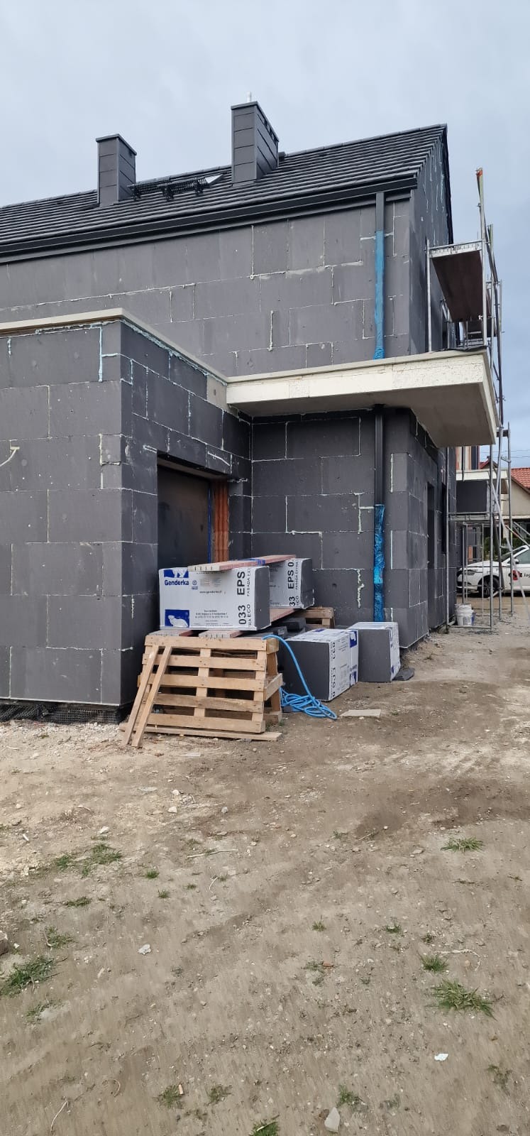 Surowy budynek w trakcie ocieplania, widoczne szare bloki izolacyjne EPS, paleta i opakowania materiału, rusztowanie z boku, dach z ciemnej dachówki.