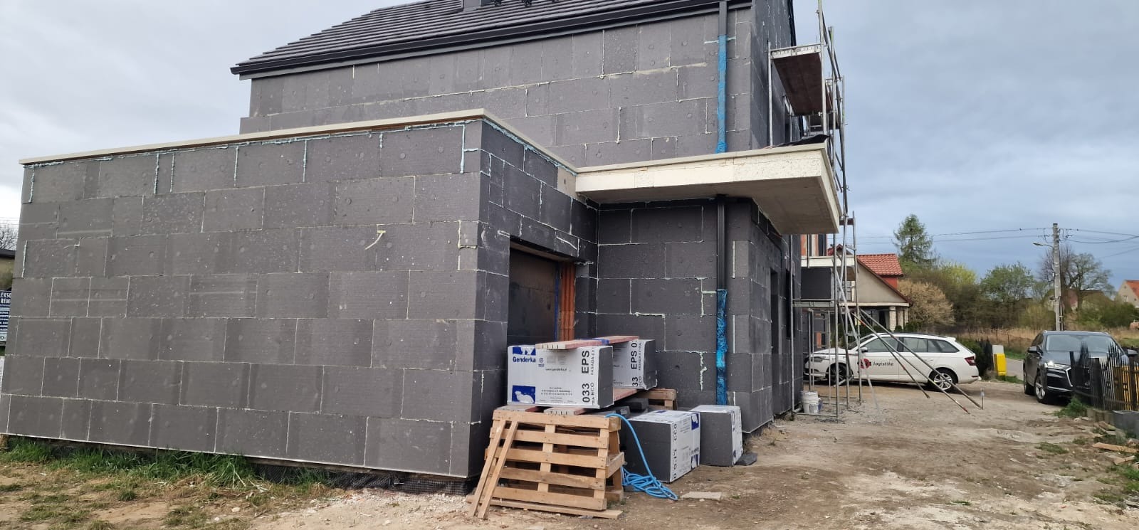 Dom w trakcie ocieplania elewacji styropianem grafitowym, widoczne płyty styropianowe, rusztowania, paleta z paczkami styropianu, przygotowane do dalszych prac.
