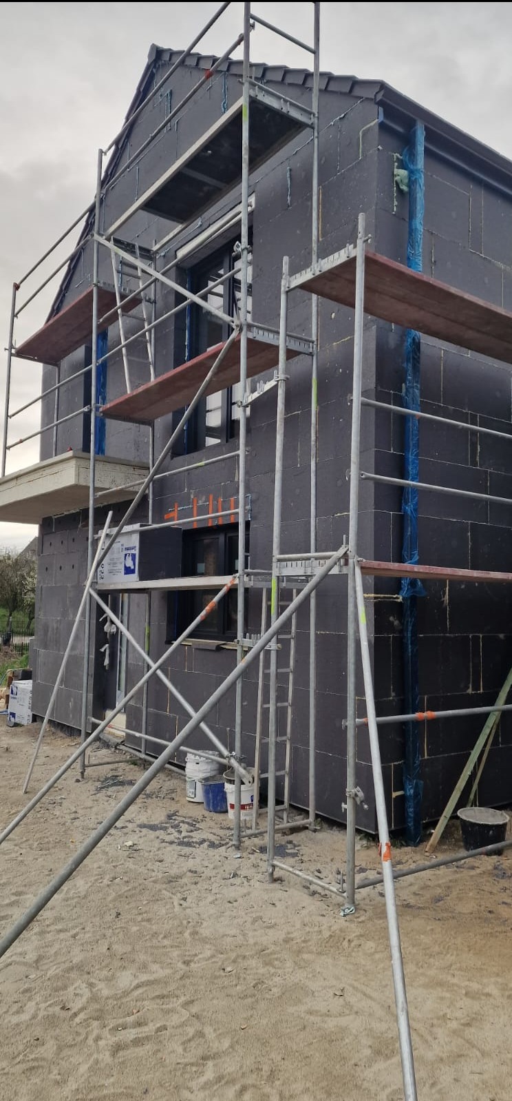 Dom w trakcie ocieplania, ściany pokryte grafitowym styropianem, widoczne rusztowania, farby i oznaczenia okien pomarańczową taśmą.