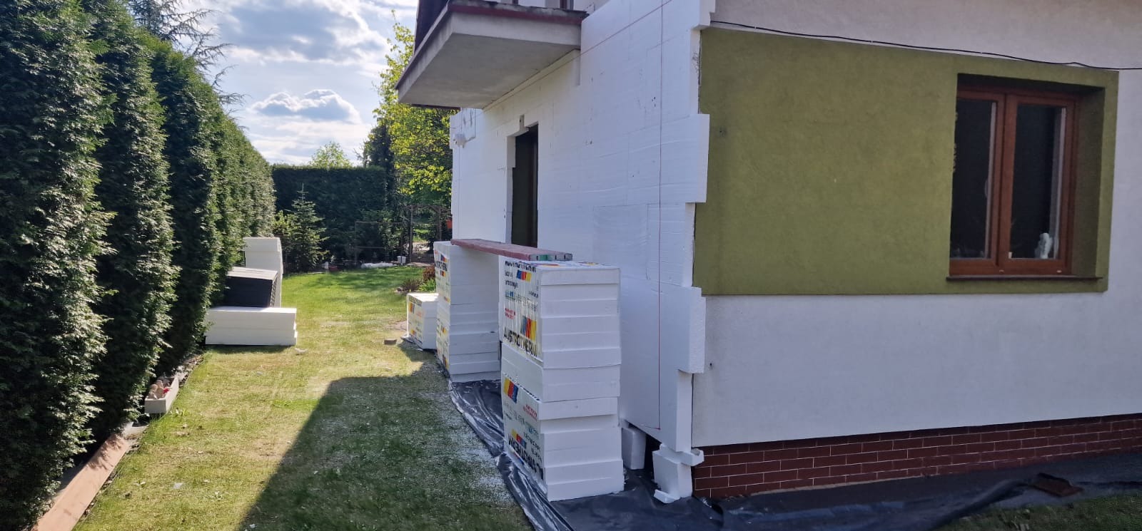 Ocieplanie domu styropianem: białe płyty izolacyjne ułożone przy ścianie, widoczne fragmenty elewacji w kolorze oliwkowym i białym, zielony trawnik i wysokie tuje w tle.