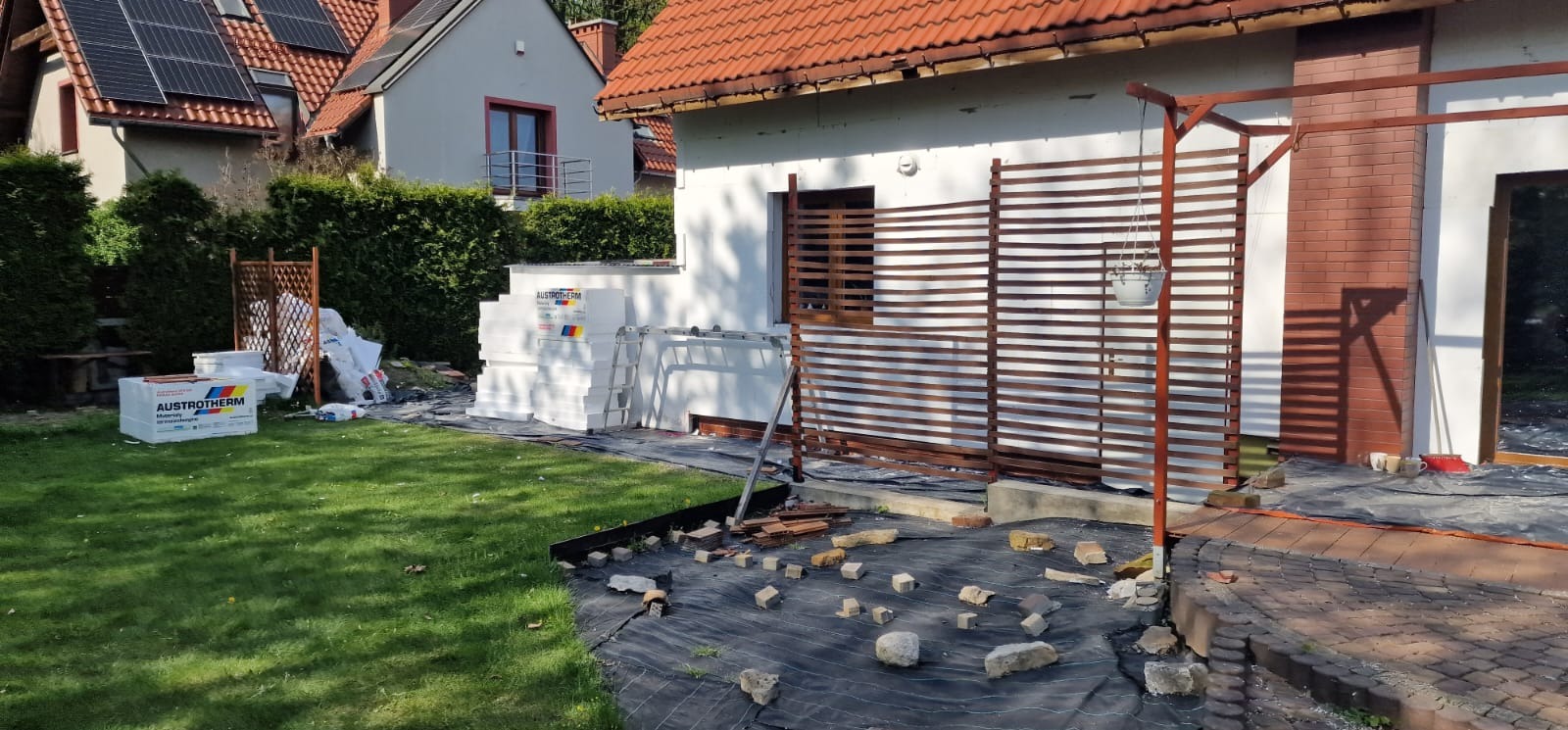 Ocieplanie elewacji domu jednorodzinnego w trakcie: styropian, rusztowanie drewniane, teren przygotowany do prac. Widok z zewnątrz, w tle dom z panelami słonecznymi.