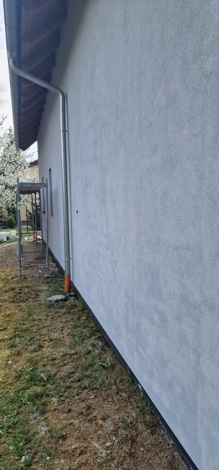 Świeżo otynkowana elewacja domu w odcieniach szarości, widoczna z boku, z rynną i fragmentem rusztowania. Trawa i drzewo w tle.