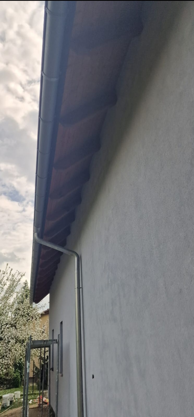 Elewacja budynku po ociepleniu z zamontowanym systemem rynnowym. Widok z dołu na szarą ścianę, rynnę i fragment dachu. W tle drzewo i błękitne niebo.