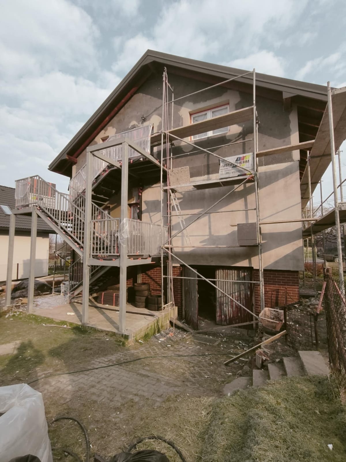Dom w trakcie ocieplania z rusztowaniem i nową klatką schodową z metalu. Widoczne materiały budowlane, elewacja w trakcie prac. Dzień, pochmurne niebo.