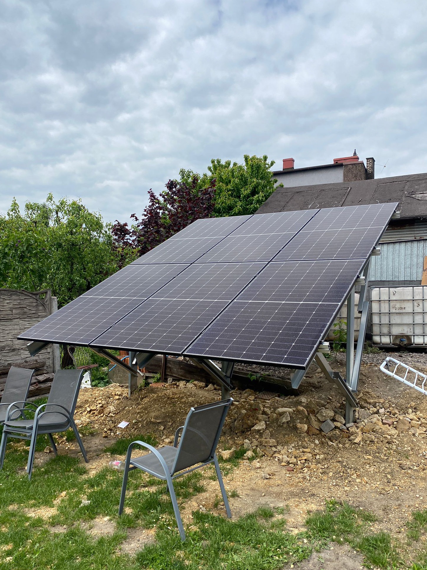 Panel słoneczny na metalowej konstrukcji w ogrodzie, widok z dołu. W tle dom i drzewa. Słoneczny dzień.