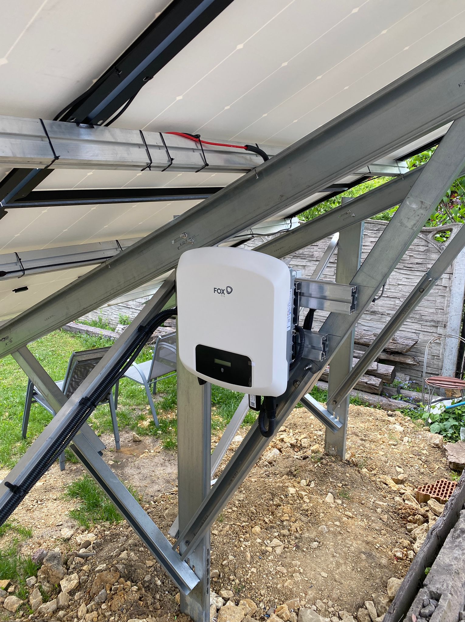 Biały falownik FOX-ESS zamontowany na metalowej konstrukcji wsporczej paneli fotowoltaicznych. Widok z dołu, na tle ziemi i zieleni.