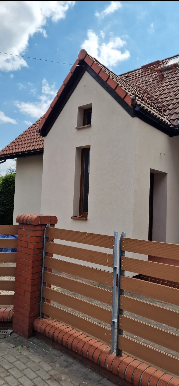 Dom po ociepleniu elewacji, z nową, jasną elewacją i drewnopodobnym ogrodzeniem. Widoczne okna i dach z czerwonej dachówki. Słoneczny dzień, błękitne niebo z chmurami.