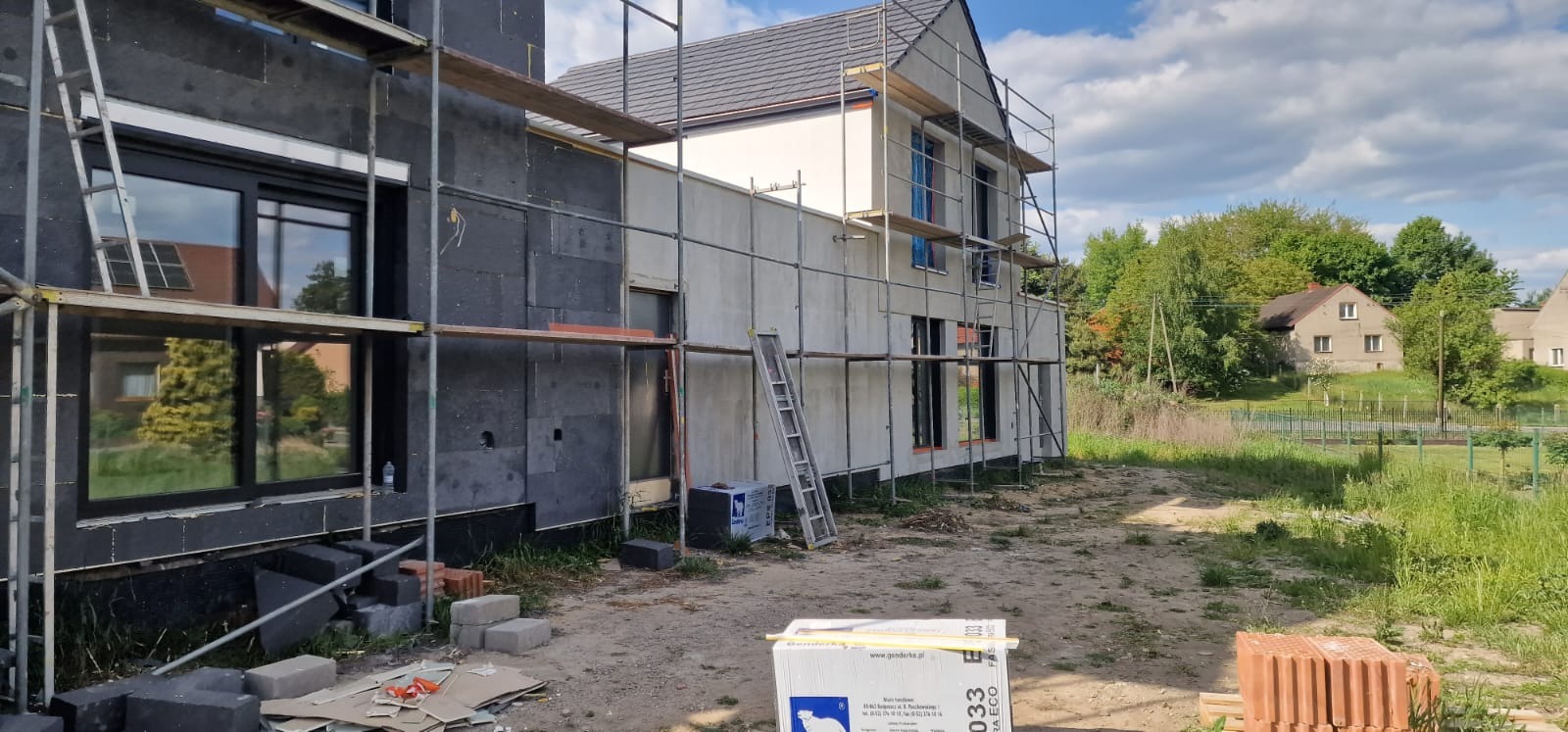 Dom w trakcie ocieplania: czarny styropian na jednej ścianie, surowa elewacja na drugiej, rusztowania, drabina, materiały budowlane na ziemi.