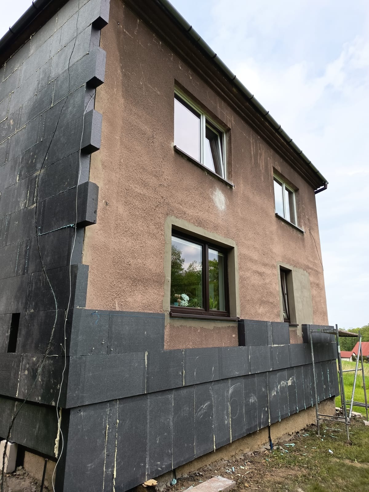 Ocieplanie elewacji budynku z widocznym styropianem grafitowym. Częściowo ocieplona ściana, widoczne okna i rusztowanie. Prace w toku, elewacja w trakcie ocieplania.