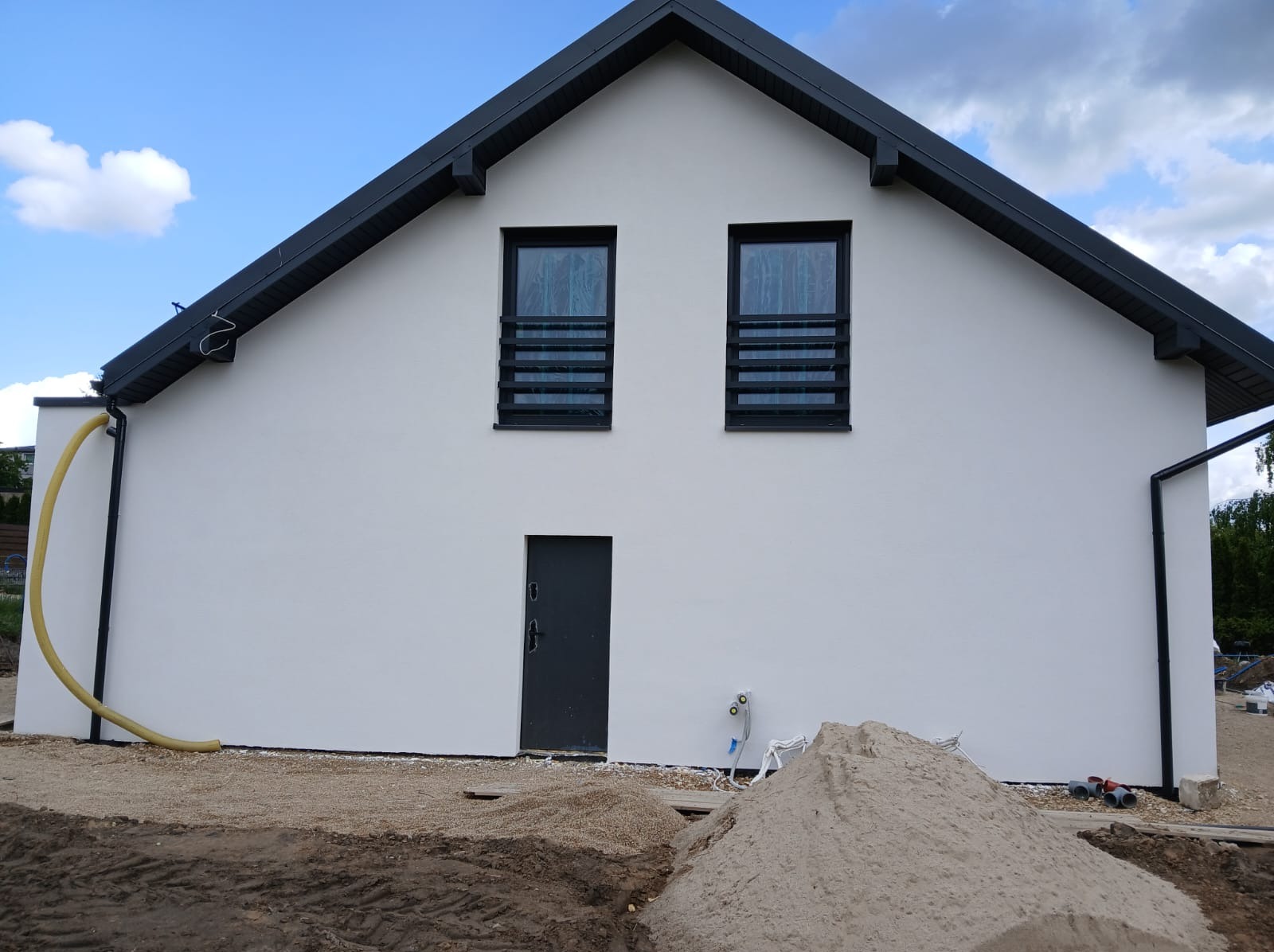 Biała elewacja domu jednorodzinnego z czarną stolarką okienną i dachem. Widoczny fragment rury i piasek sugerują prace wykończeniowe.