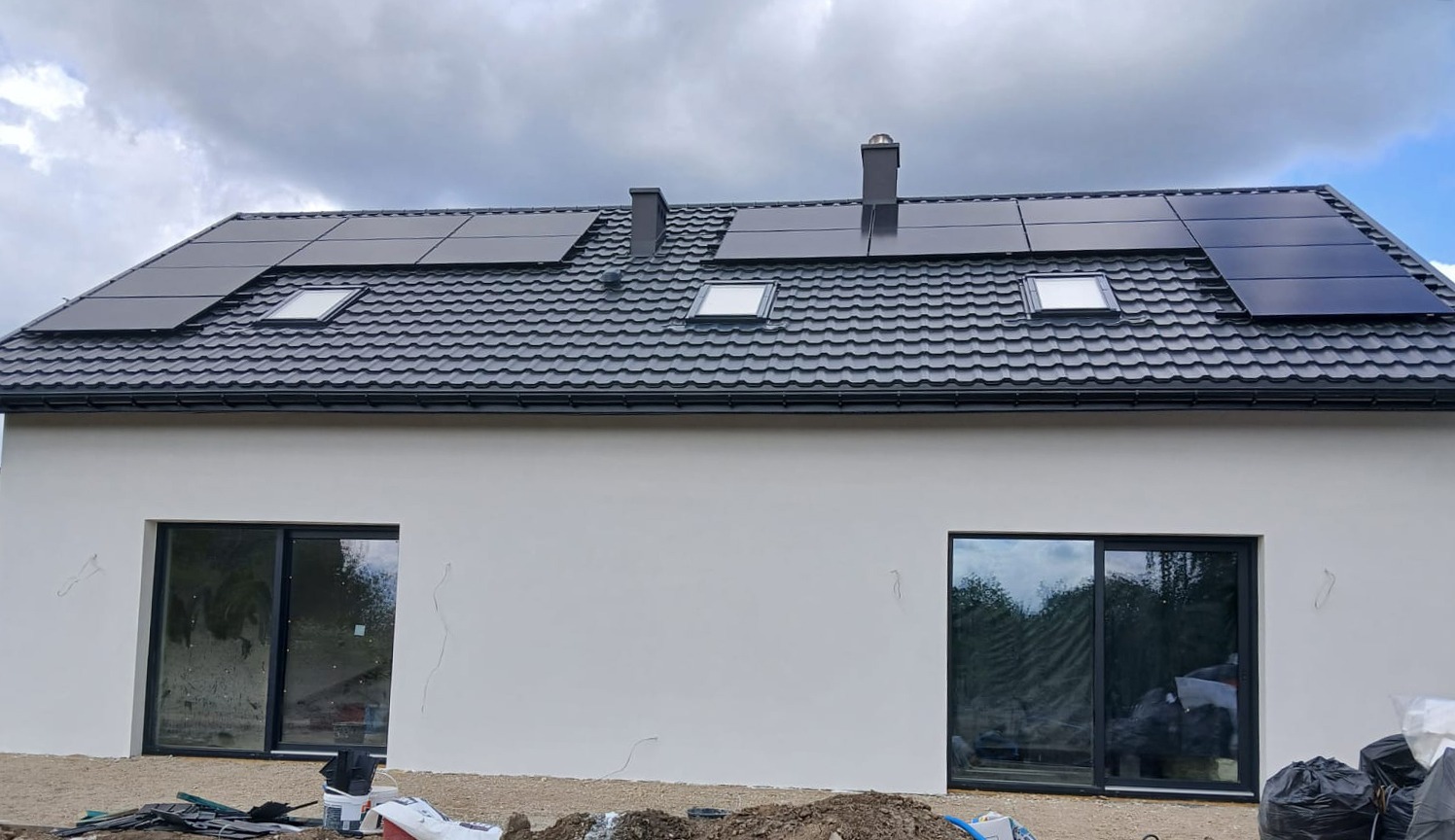 Dom jednorodzinny z panelami fotowoltaicznymi na dachu, widoczne okna dachowe i świeżo otynkowana elewacja. Ujęcie z poziomu gruntu, pochmurne niebo w tle.