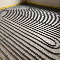 Szare rury ogrzewania podłogowego, równolegle ułożone na izolacji z czerwoną siatką, przymocowane niebieskimi uchwytami. Żółta taśma brzegowa wzdłuż ścian.