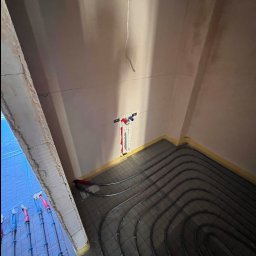 ADMAR INSTALACJE HYDRAULIKA - Wnętrze w trakcie montażu ogrzewania podłogowego. Rury grzewcze ułożone na izolacji, widoczne przyłącza do instalacji. Żółta taśma zabezpieczająca przy ścianach.