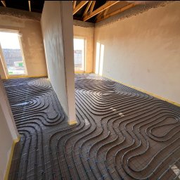 ADMAR INSTALACJE HYDRAULIKA - Rozległy widok na ułożone rury ogrzewania podłogowego w surowym wnętrzu z oknami i widoczną konstrukcją dachu. Równoległe pętle rur na folii, przygotowane do zalania.