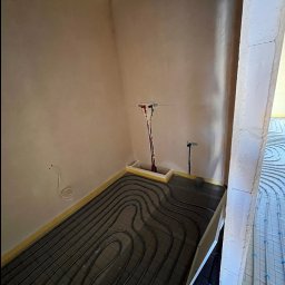 ADMAR INSTALACJE HYDRAULIKA - Ogrzewanie podłogowe w Śremie: rury instalacji ułożone na czarnej folii, widoczne wyprowadzenia instalacji na ścianie. Przygotowanie do wylewki.