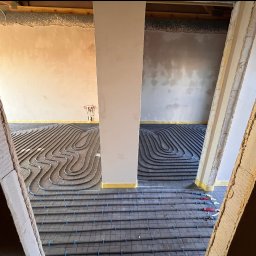 ADMAR INSTALACJE HYDRAULIKA - Ułożone rury ogrzewania podłogowego na izolacji, widoczne przez surową ramę drzwiową. Fragment ściany z instalacją hydrauliczną w tle.