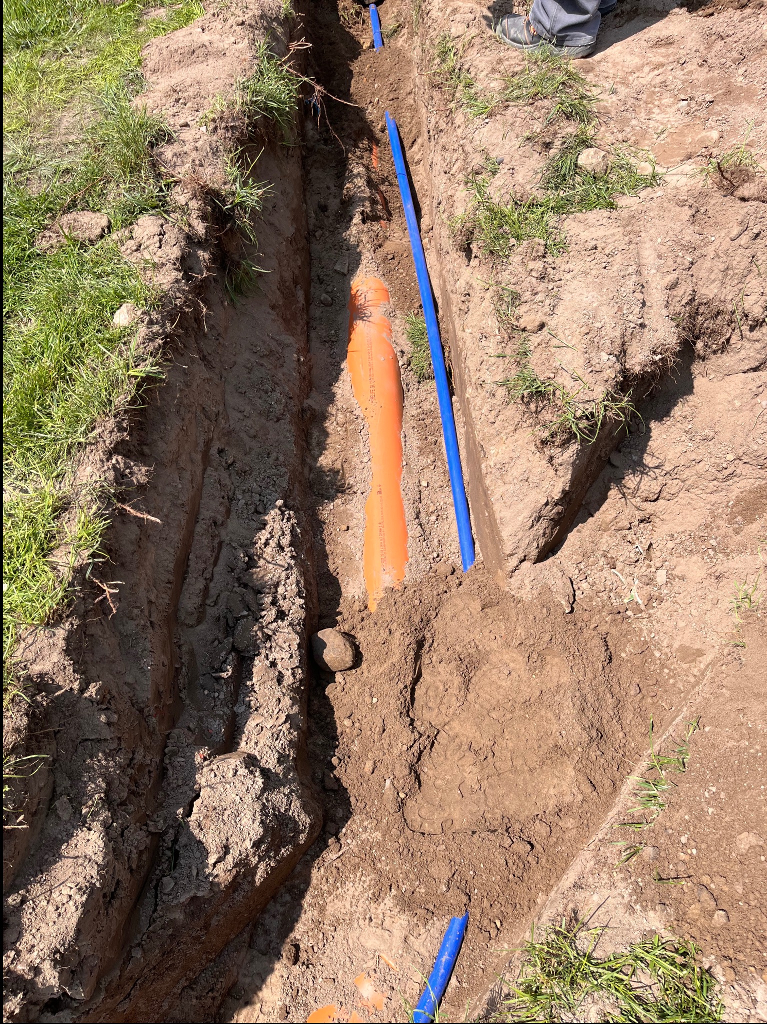 Wykop z ułożonymi rurami (pomarańczowa i niebieska) w ziemi, widoczne trawy na krawędzi wykopu, fragment buta osoby w prawym górnym rogu. Instalacja podziemna.