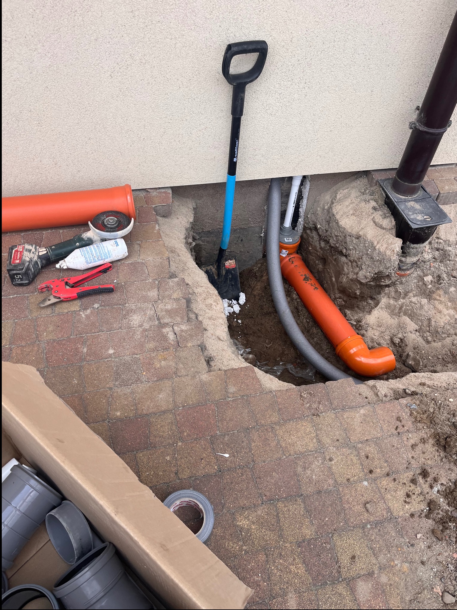 Wykop z ułożonymi rurami kanalizacyjnymi, widoczne narzędzia: szpadel, szlifierka kątowa, nożyce do rur. Pomarańczowe rury kanalizacyjne w wykopie przy ścianie budynku.