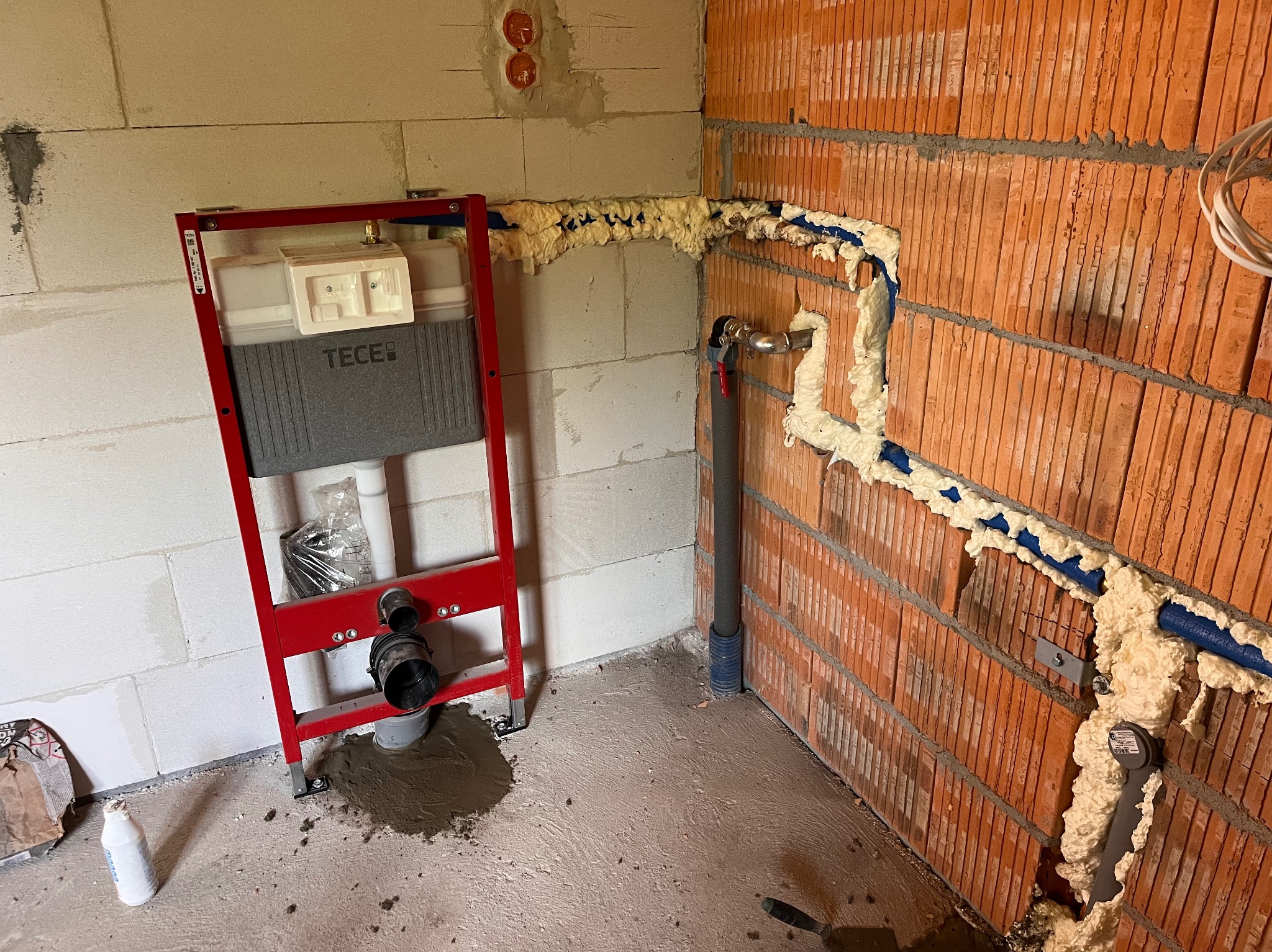 Stelaż podtynkowy TECE w czerwonej ramie, przygotowany do montażu. Widoczne niebieskie rury instalacyjne zabezpieczone pianką montażową na ścianie z cegły.