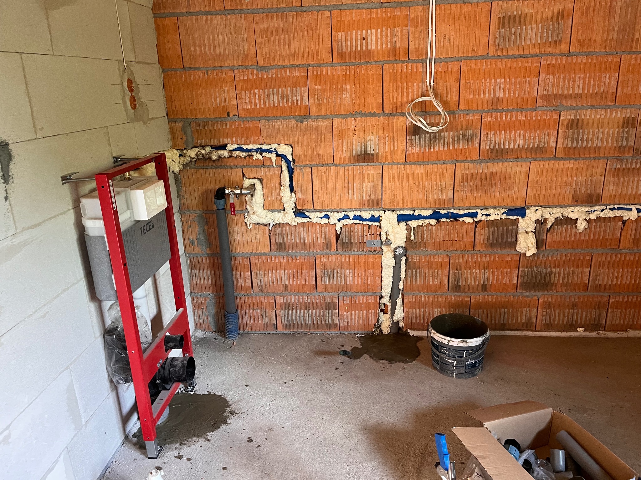 Niezabudowana instalacja podtynkowa WC z rurami, zabezpieczona pianką montażową, na tle ściany z czerwonej cegły dziurawki. Widoczne elementy montażowe i instalacyjne.