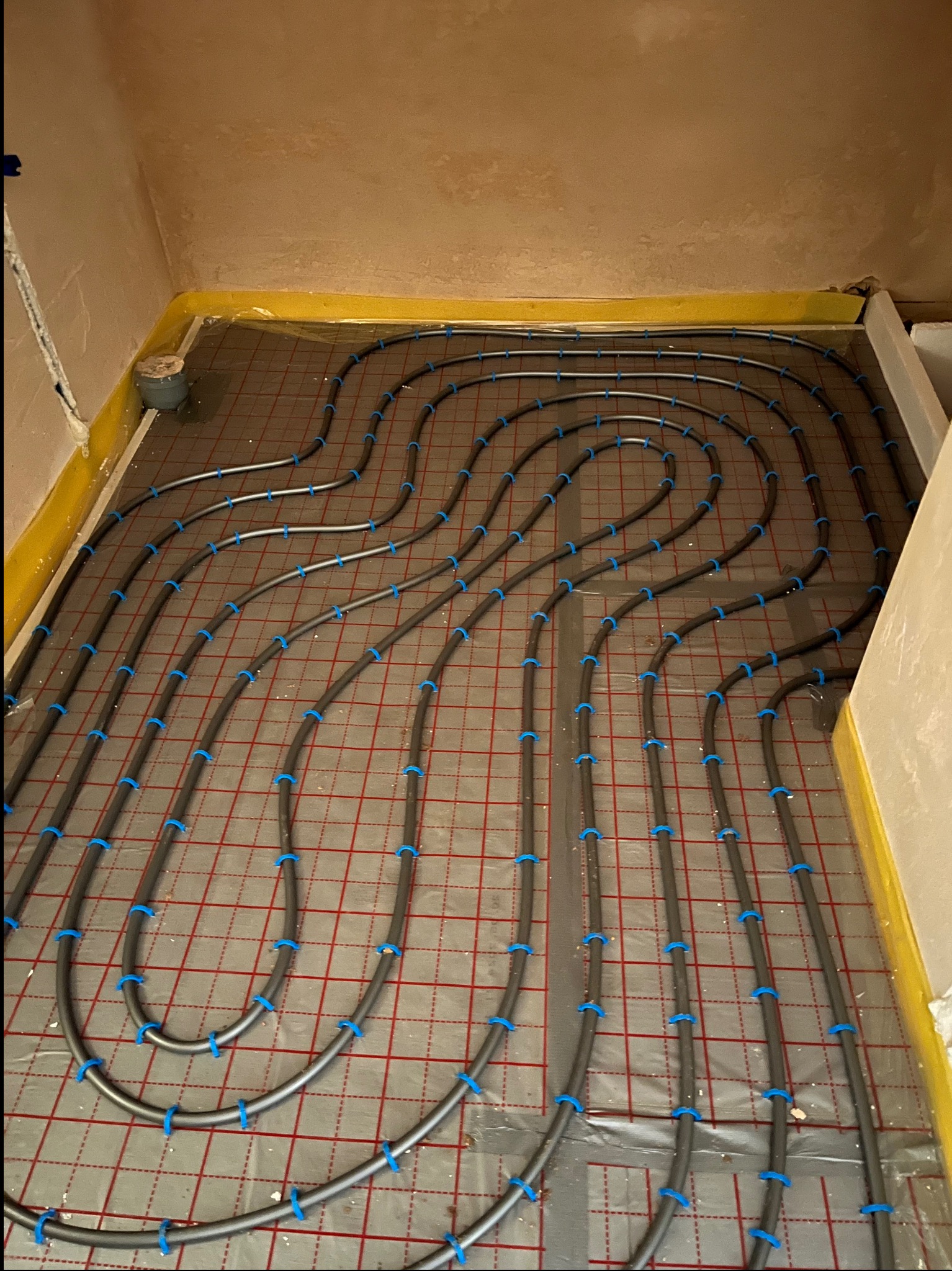 Szare rury ogrzewania podłogowego, mocowane niebieskimi uchwytami do folii z czerwoną siatką, ułożone w pętlach na przygotowanym podłożu w pomieszczeniu.