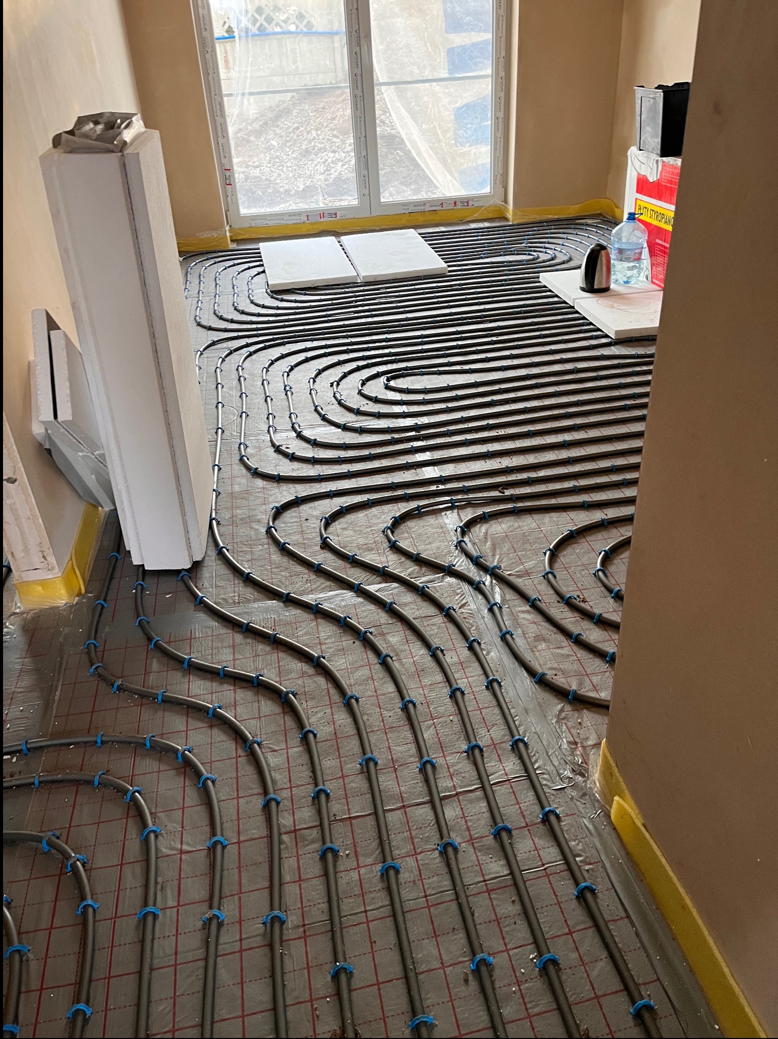 Ułożone rury ogrzewania podłogowego na folii z siatką w nowym pomieszczeniu, widoczne okno z zewnątrz i materiały budowlane. Charakterystyczne niebieskie uchwyty mocujące.