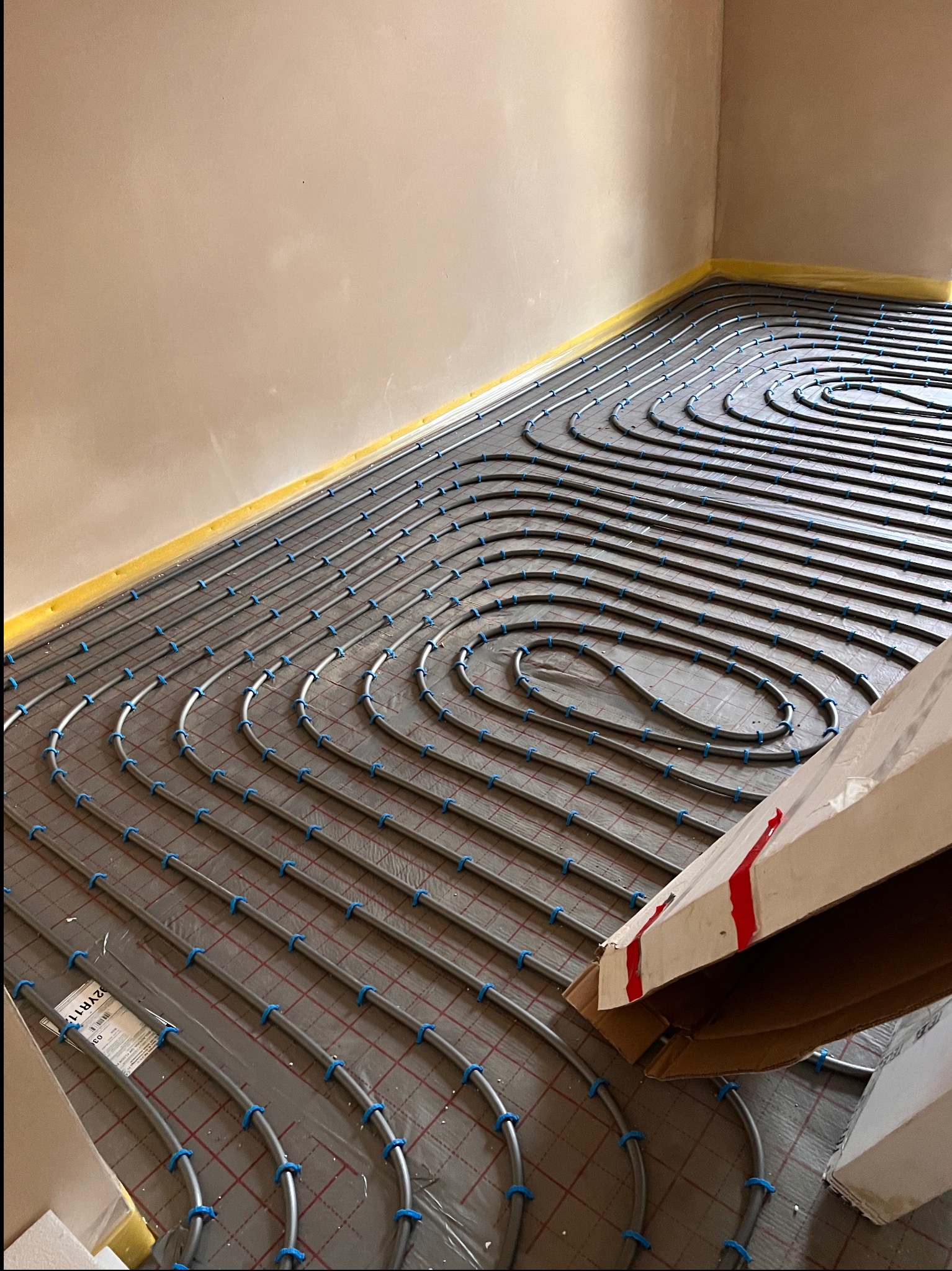 Szare rury ogrzewania podłogowego, mocowane niebieskimi uchwytami do podłoża z siatką. Widoczne kartony i zabezpieczenia ścian folią. Ujęcie z góry, ukośne.