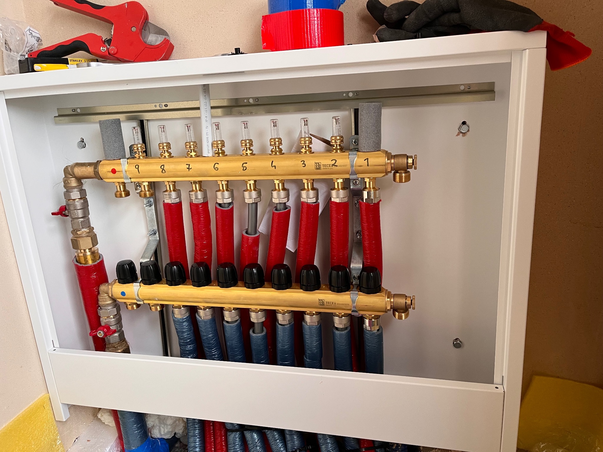 Rozdzielacz ogrzewania podłogowego Tece w białej szafce, z numerowanymi sekcjami od 1 do 9, z czerwonymi i niebieskimi rurami, narzędzia na górnej półce.