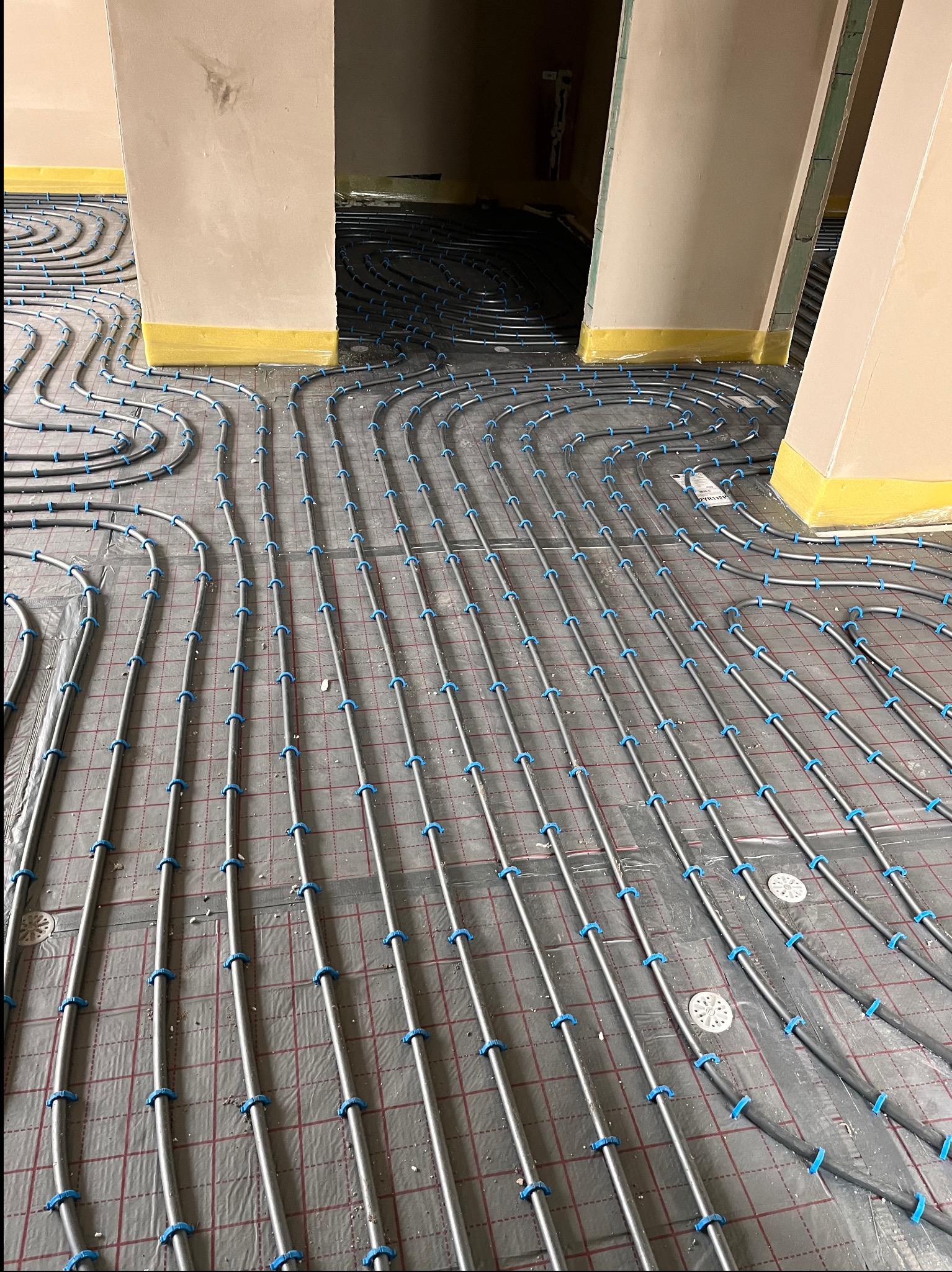Ułożone rury ogrzewania podłogowego na folii z rastrem, mocowane niebieskimi klipsami. Widoczne fragmenty ścian z żółtą taśmą izolacyjną przy podłodze.