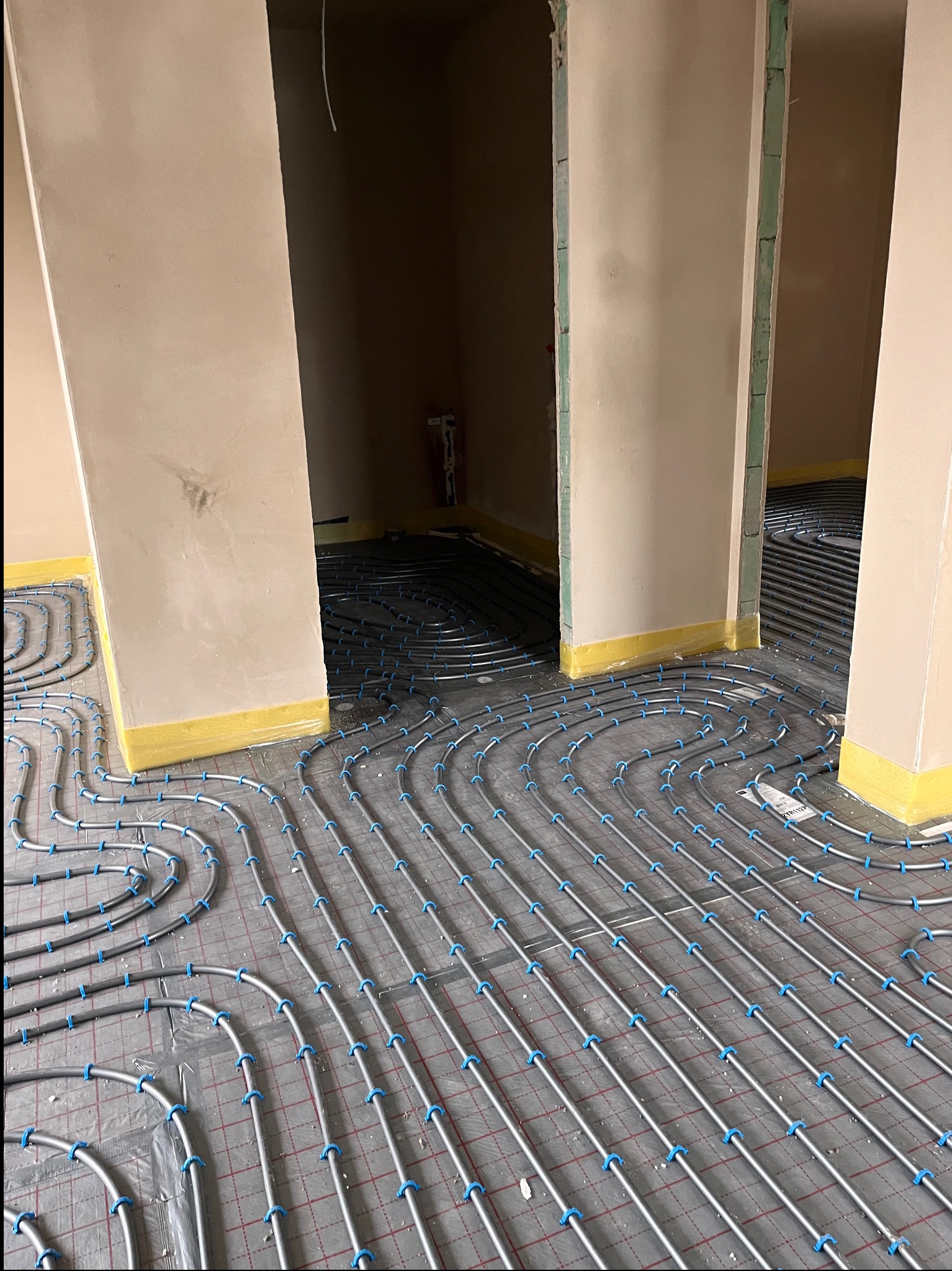 Szare rury ogrzewania podłogowego z niebieskimi uchwytami ułożone na czarnej folii z czerwoną kratką, widoczne fragmenty ścian z żółtą taśmą.