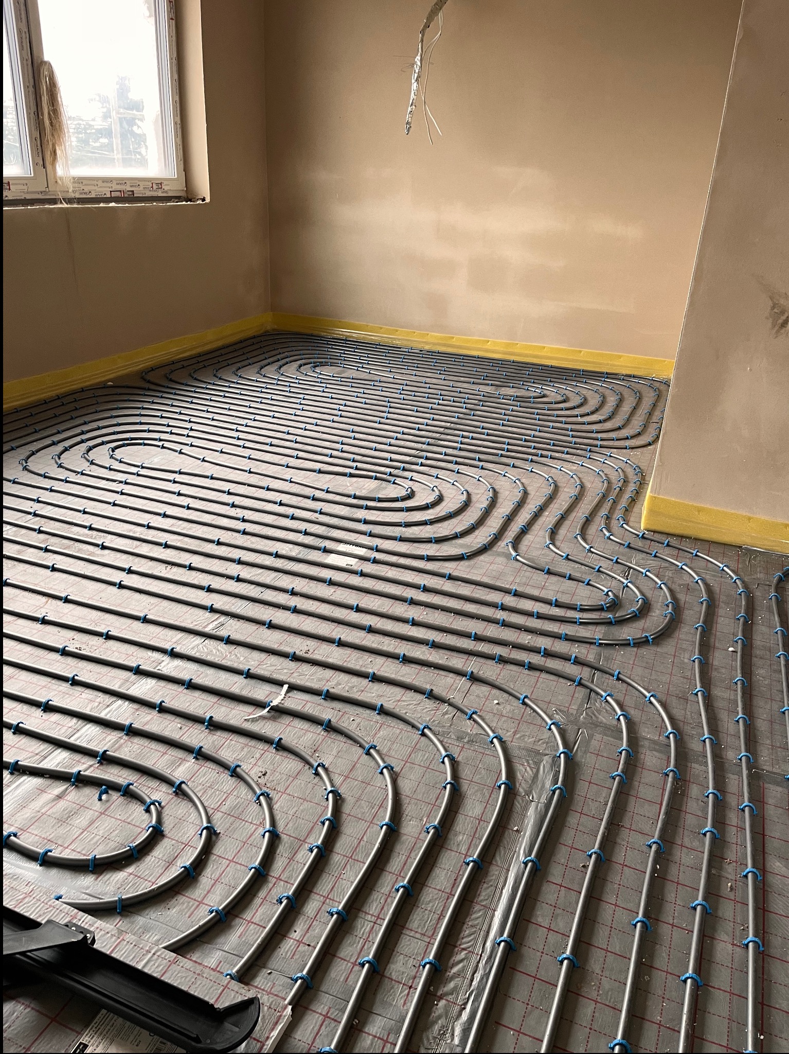 Szare rury ogrzewania podłogowego, z niebieskimi uchwytami, ułożone na folii z siatką w surowym pomieszczeniu z oknem. Żółta taśma przy ścianach.
