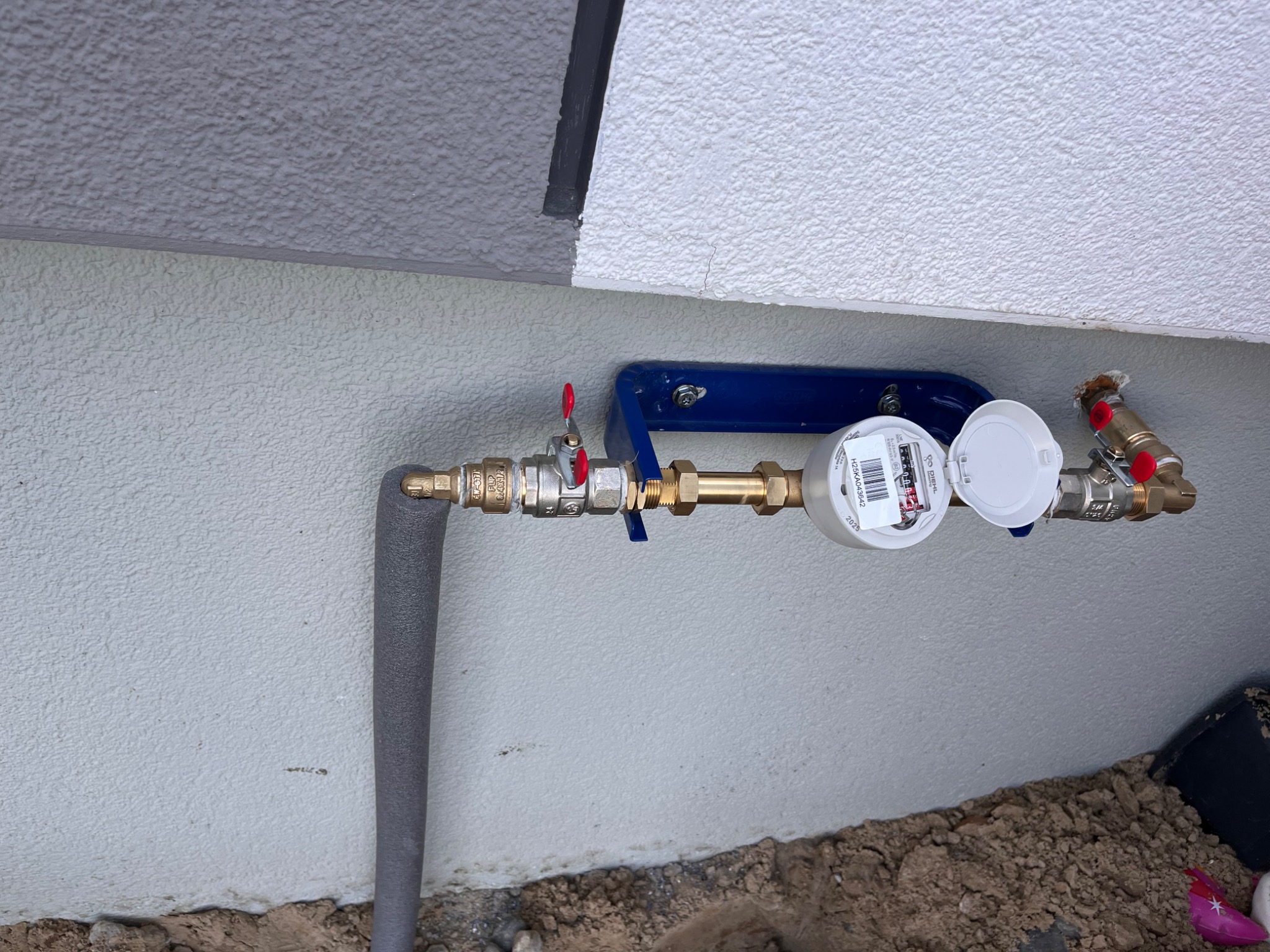 Zamontowany wodomierz na zewnątrz budynku, z widoczną izolacją rury i elementami armatury. Ujęcie z bliska, ukazujące szczegóły instalacji przy ścianie.